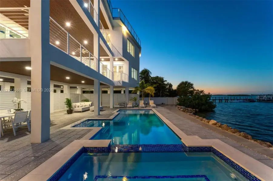 Waterfront multi-level residence featuring a custom swimming pool with integrated spa, expansive covered lanais, metal cable railings, and textured stone-finish decking Waterfront multi-level residence featuring a custom swimming pool with integrated spa, expansive covered lanais, metal cable railings, and textured stone-finish decking