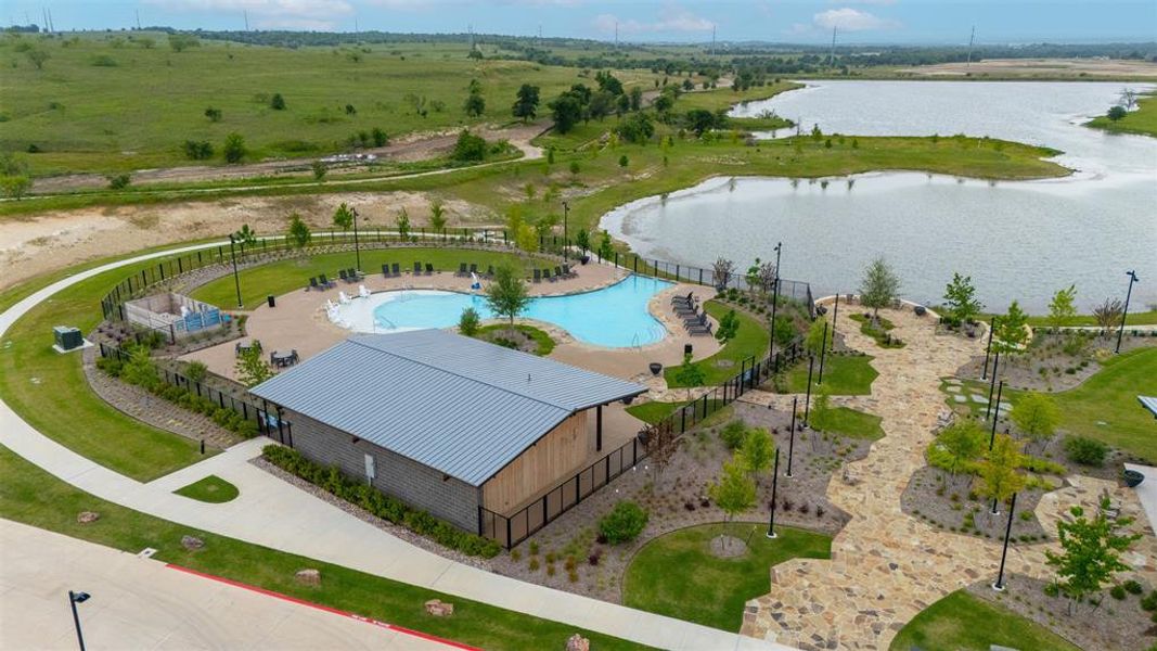Recreational amenity featuring a community swimming pool, an open-air pavilion with a metal roof, a stone-paved pathway, and a surrounding landscaped lawn area, all bordering a natural body of water