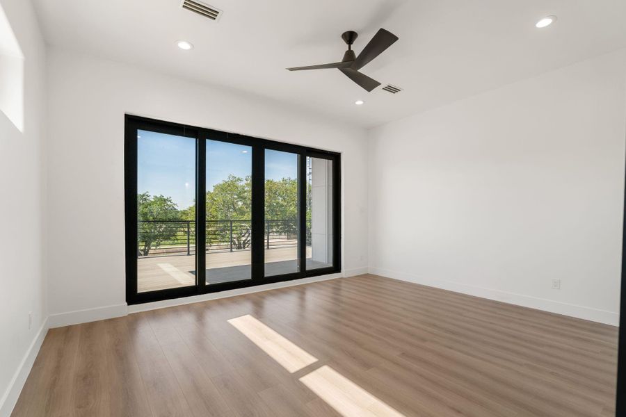 This upstairs secondary bedroom opens directly to the rooftop deck through a bank of large black-framed sliding glass panels, flooding the room with natural light and giving it a direct outdoor connection that most bedrooms never have. Light vinyl plank flooring and a dark-blade ceiling fan carry the home's contemporary palette through every detail.