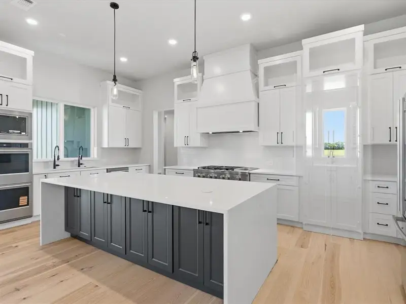 Gourmet kitchen featuring a large central island with dark cabinetry, light wood-finish flooring, white perimeter cabinetry, stainless steel appliances, and quartz countertops