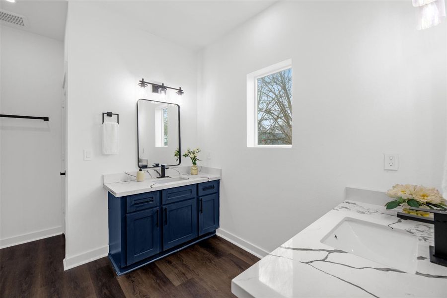 Lovely modern bathroom with a stylish blue vanity, marble countertop, and sleek fixtures. Features a large mirror, natural light from a window, and elegant dark wood flooring.