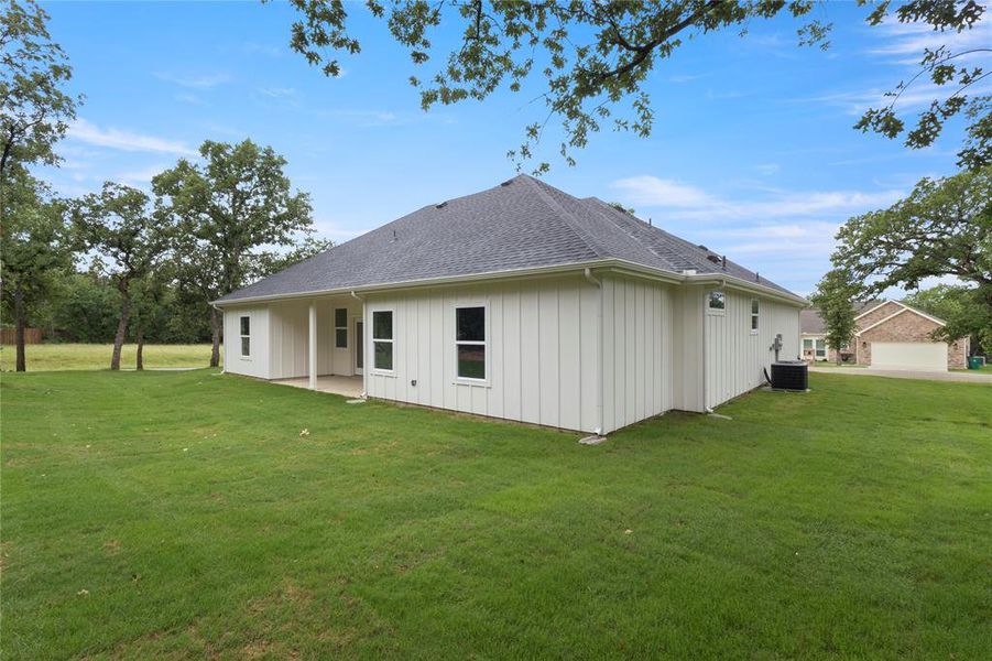 Expansive green lawn surrounds this property featuring white vertical siding, a dark shingle roof, and a covered concrete patio Expansive green lawn surrounds this property featuring white vertical siding, a dark shingle roof, and a covered concrete patio