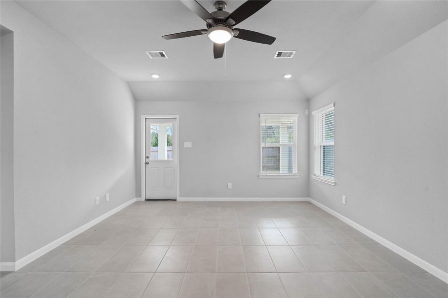 A bright and airy open-concept space featuring tile flooring, sloped ceilings with recessed lighting and ceiling fan, abundant natural light from large windows (fitted with blinds), and an access door to the back covered patio.