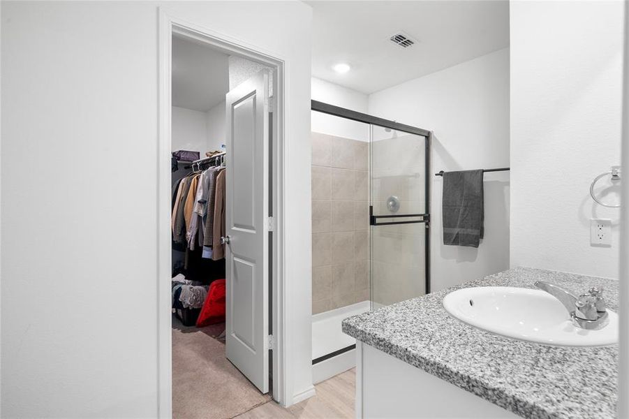 Bathroom with a single vanity featuring a granite-look countertop and an integrated sink, a glass-enclosed shower with tiled walls, and a walk-in closet with carpet, and walk in show featuring glass doors Bathroom with a single vanity featuring a granite-look countertop and an integrated sink, a glass-enclosed shower with tiled walls, and a walk-in closet with carpet, and walk in show featuring glass doors