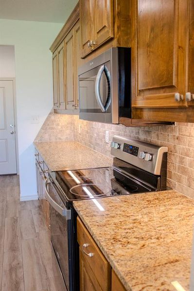 Kitchen with appliances with stainless steel finishes, backsplash, light wood finished floors, brown cabinetry, and light stone counters Kitchen with appliances with stainless steel finishes, backsplash, light wood finished floors, brown cabinetry, and light stone counters
