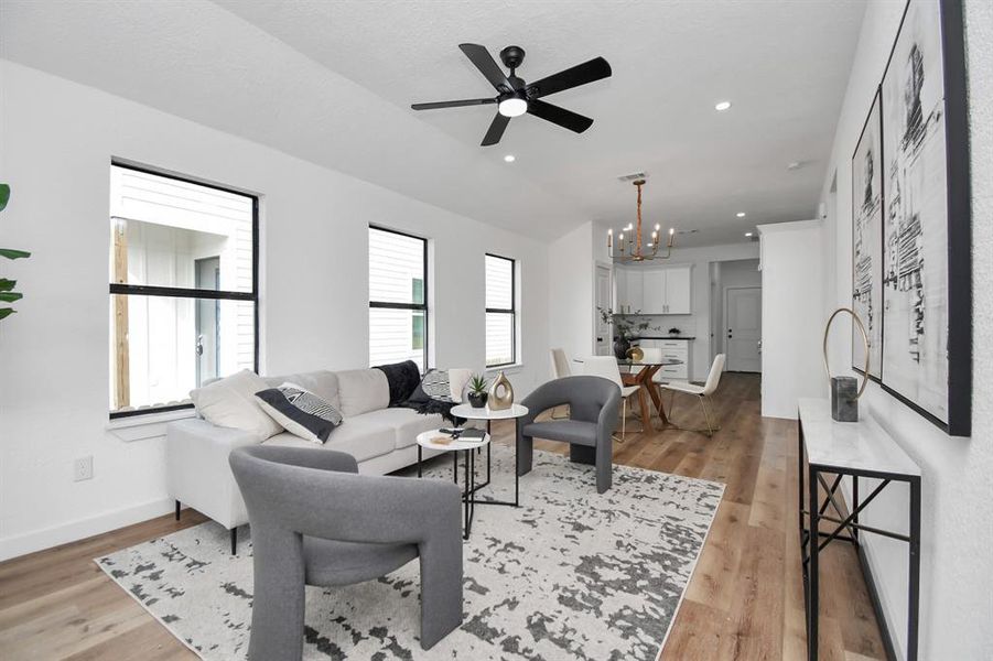 This is a modern and bright living room featuring high ceilings with a ceiling fan, hardwood floors, contemporary furnishings, and large windows that let in plenty of natural light. The room is styled with a neutral color palette and includes decorative touches such as wall art, plants, and a patterned area rug.