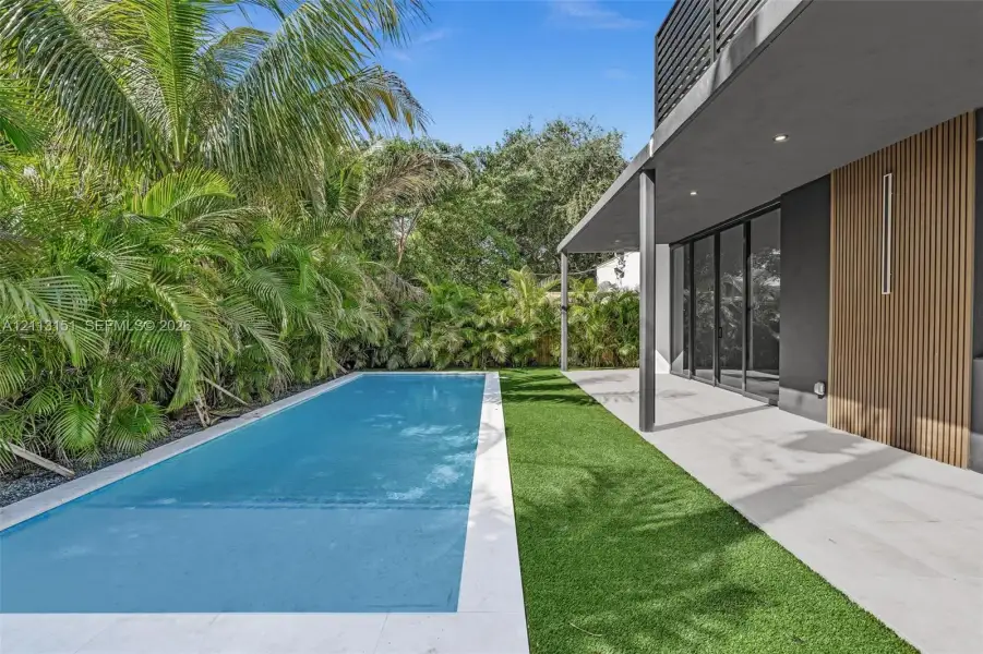 Rectangular swimming pool featuring a light-toned stone surround, synthetic lawn, covered patio with recessed lighting, and exterior wood-slat accent wall Rectangular swimming pool featuring a light-toned stone surround, synthetic lawn, covered patio with recessed lighting, and exterior wood-slat accent wall