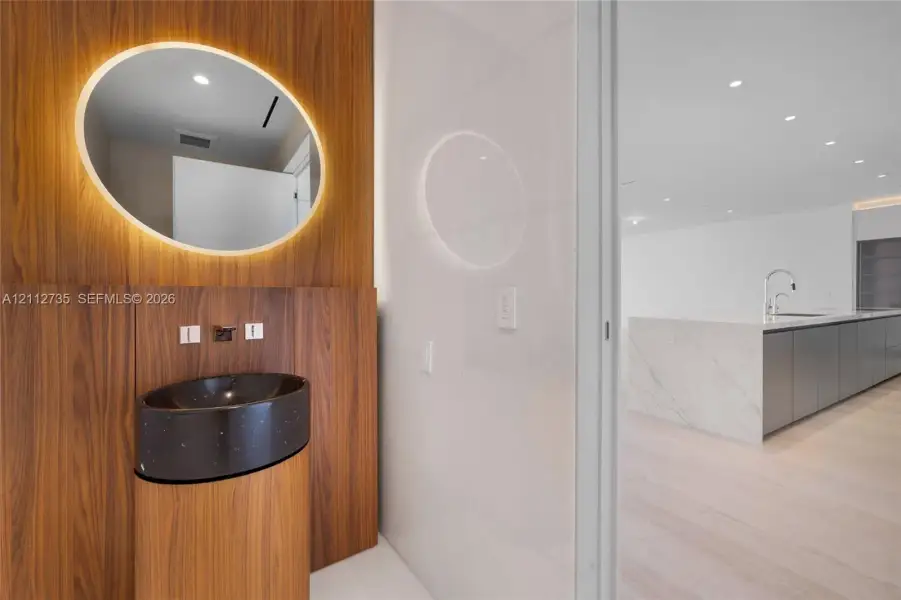 Wood-paneled wall feature with integrated backlit oval mirror, dark speckled pedestal basin, wall-mounted faucet, and stone-surfaced kitchen island with matte cabinetry