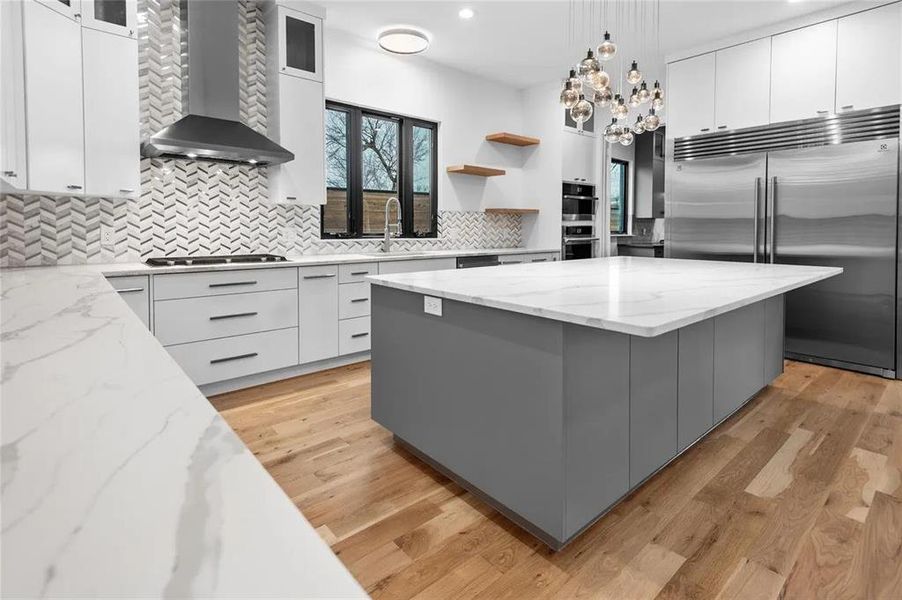 Contemporary kitchen featuring a spacious center island with a white countertop, stainless steel appliances, herringbone pattern backsplash, light wood-finish flooring, and minimalist cabinetry Contemporary kitchen featuring a spacious center island with a white countertop, stainless steel appliances, herringbone pattern backsplash, light wood-finish flooring, and minimalist cabinetry