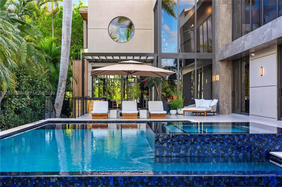 Oversized pool and elevated spa with blue mosaic tile waterfall spillover, teak chaise lounges, cantilever umbrella, woven lounge seating, large-format stone pool deck, floor-to-ceiling glass rear elevation, stone cladding, circular porthole window, and glass-enclosed wellness center beyond.