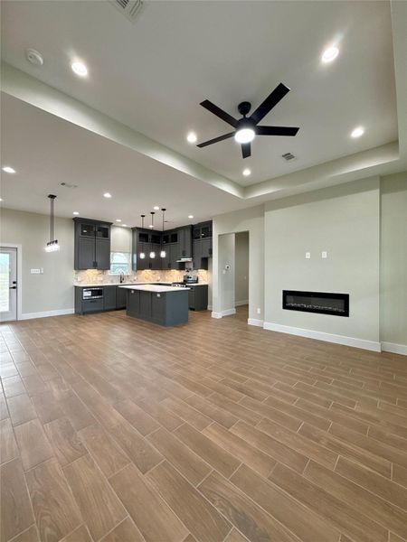 This modern, open concept living area features a sleek kitchen with dark cabinetry, a large island, and elegant pendant lighting. The space is accented by wood-look tile flooring, recessed lighting, and a contemporary ceiling fan. A built-in elegant wall fireplace adds warmth and style.