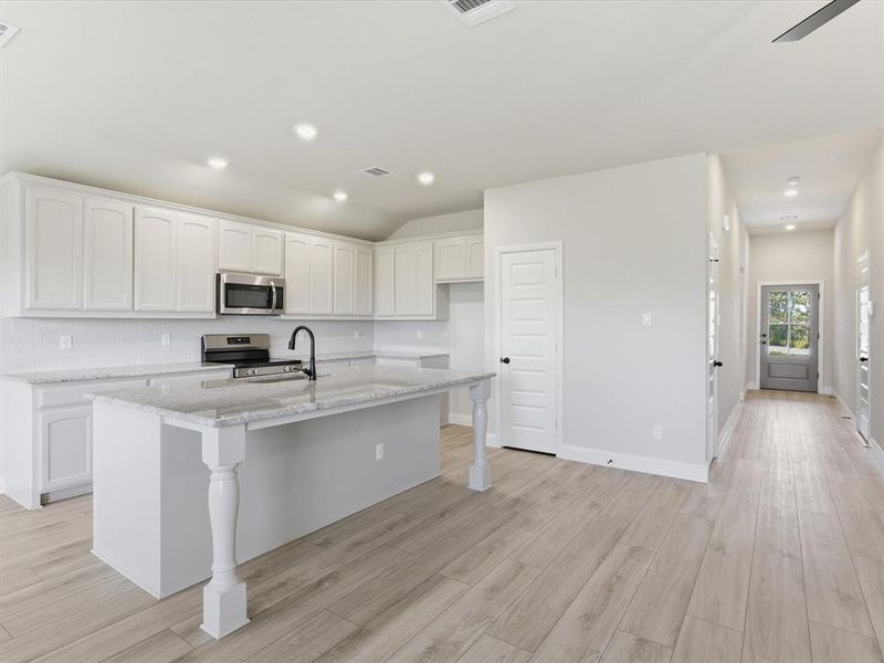 Open-concept kitchen with light wood-finish flooring, white cabinetry, a large central island featuring decorative supports, integrated stainless steel appliances, and recessed lighting Open-concept kitchen with light wood-finish flooring, white cabinetry, a large central island featuring decorative supports, integrated stainless steel appliances, and recessed lighting
