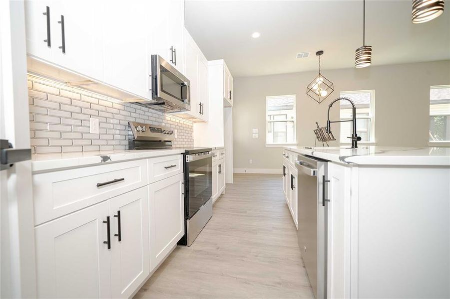 Kitchen with appliances with stainless steel finishes, white cabinetry, backsplash, light stone counters, and pendant lighting Kitchen with appliances with stainless steel finishes, white cabinetry, backsplash, light stone counters, and pendant lighting