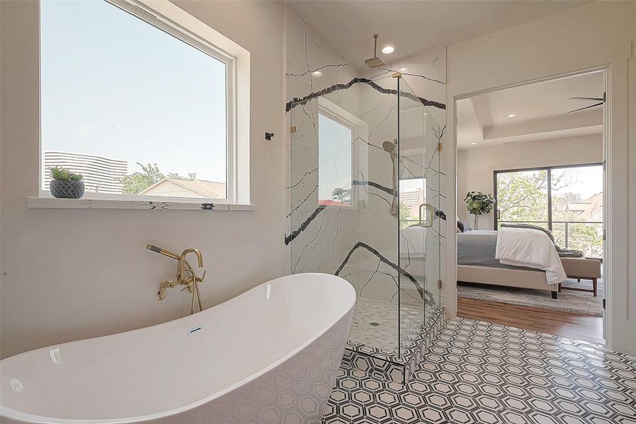 This is a modern bathroom with a freestanding tub, gold fixtures, a large window for natural light, and a glass shower enclosure with a unique marble design. The room connects to a bedroom with a view of the outdoors. This is a modern bathroom with a freestanding tub, gold fixtures, a large window for natural light, and a glass shower enclosure with a unique marble design. The room connects to a bedroom with a view of the outdoors.