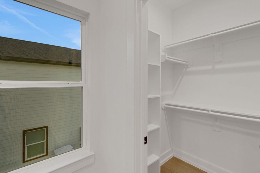 This photo shows a bright, walk-in closet with ample shelving and hanging space. It features a window providing natural light and has a clean, modern design with white walls and light wood flooring. This photo shows a bright, walk-in closet with ample shelving and hanging space. It features a window providing natural light and has a clean, modern design with white walls and light wood flooring.