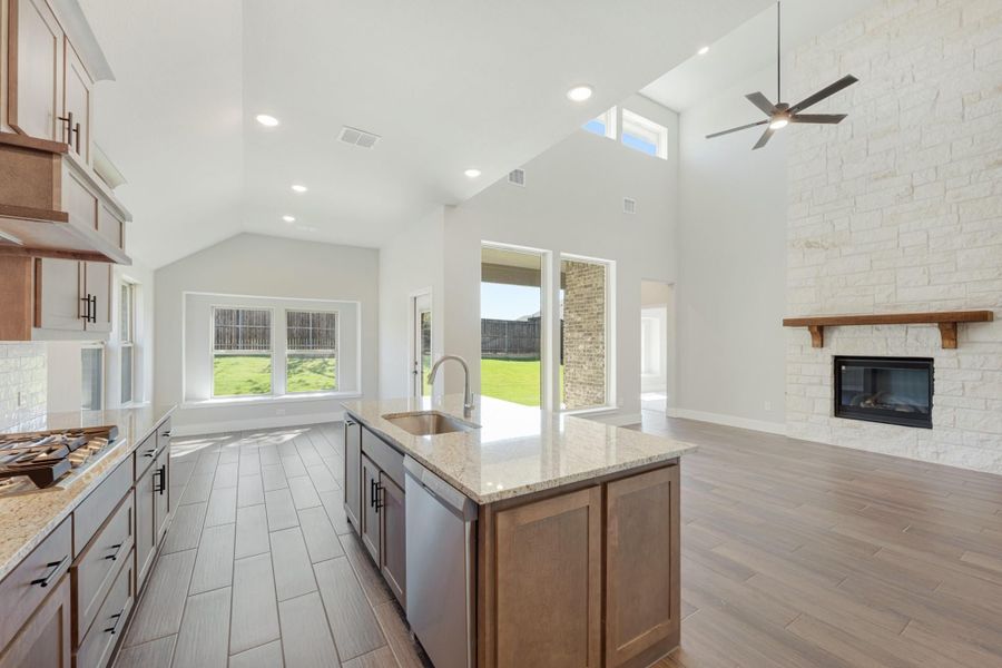 Open-concept kitchen with granite island, wood cabinets, vaulted ceiling, and stone fireplace visible in adjoining living area. Open-concept kitchen with granite island, wood cabinets, vaulted ceiling, and stone fireplace visible in adjoining living area.