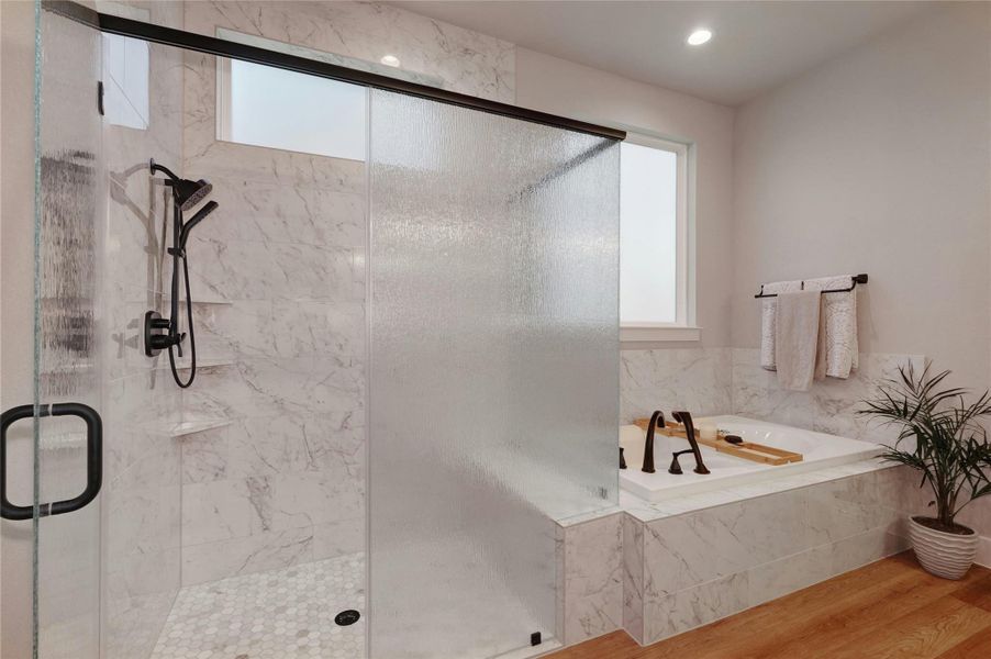 Bathroom featuring a built-in tub with a dark bronze faucet, a glass-enclosed shower with a multi-function showerhead, marble-patterned tile, and wood-finish flooring
