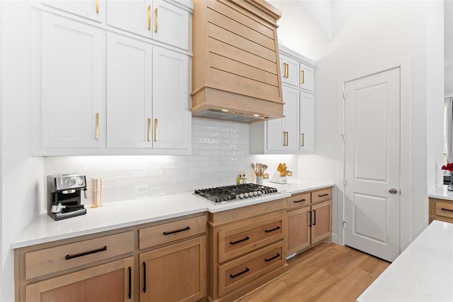 The kitchen features white upper cabinetry with gold-toned hardware, light wood-toned lower cabinetry with dark hardware, and white countertops