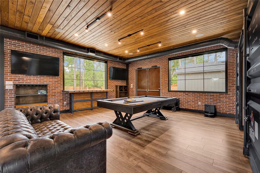 Stylish brick walls, a wooden ceiling with recessed lighting, and large windows for ample natural light. It includes a pool table, a comfortable leather sofa, and multiple wall-mounted TVs, creating an ideal space for relaxation and gatherings.