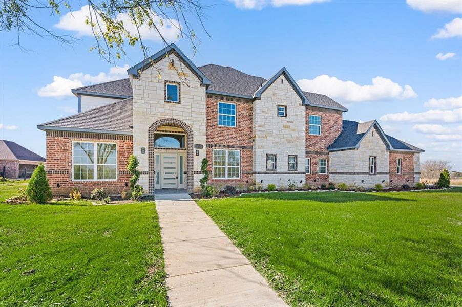 View of front facade featuring a front yard, stone siding, brick siding, and a shingled roof View of front facade featuring a front yard, stone siding, brick siding, and a shingled roof