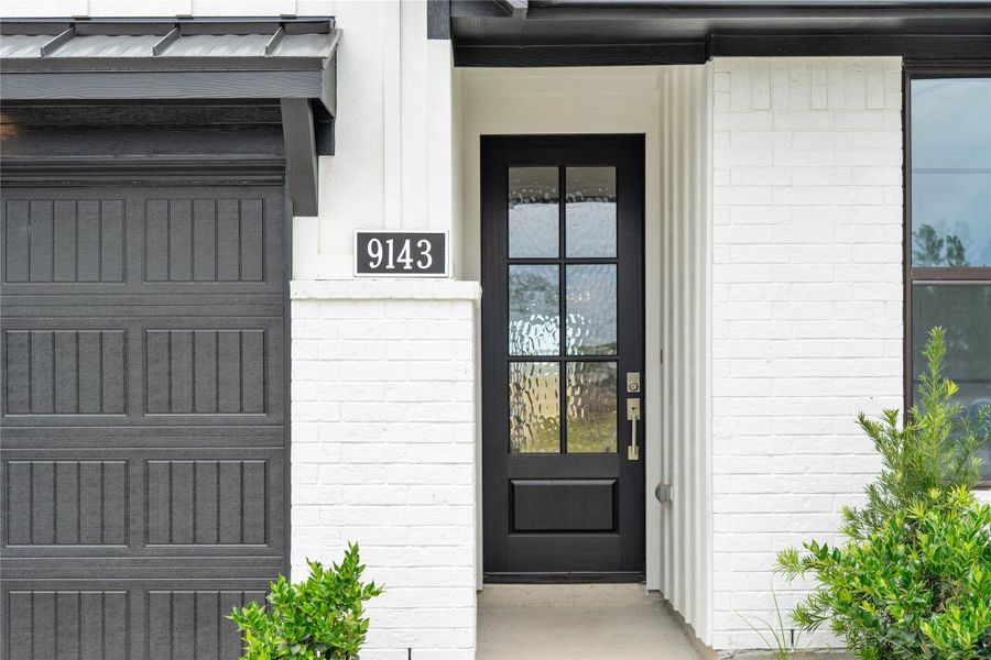 Welcome guests through a sophisticated entry door that serves as a bold focal point against the crisp, white-painted brick exterior. The door features elegant simulated divided lites and textured privacy glass, allowing natural light to fill the foyer while maintaining home security.