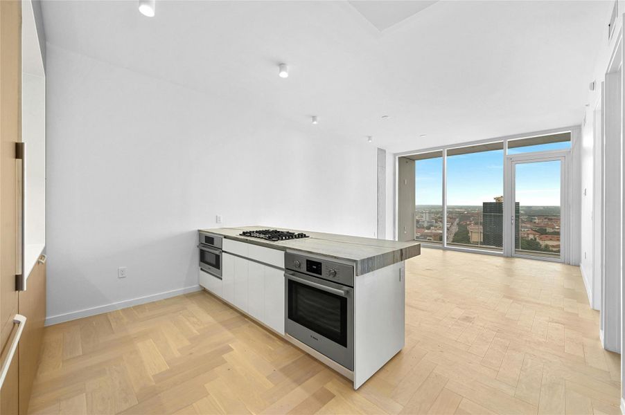 A modern, open-concept kitchen, dining and living area. The kitchen features a sleek, minimalist design with a large island at its center, topped with a light-colored stone countertop. The island includes a built-in gas cooktop and stainless steel oven. A modern, open-concept kitchen, dining and living area. The kitchen features a sleek, minimalist design with a large island at its center, topped with a light-colored stone countertop. The island includes a built-in gas cooktop and stainless steel oven.