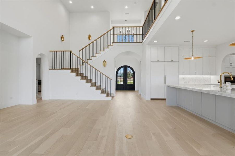 Open-concept living area featuring light wood-finish flooring, a grand staircase with black metal railings, a gourmet kitchen island with marble-look countertops, white cabinetry, and a prominent arched entryway with a dark double door