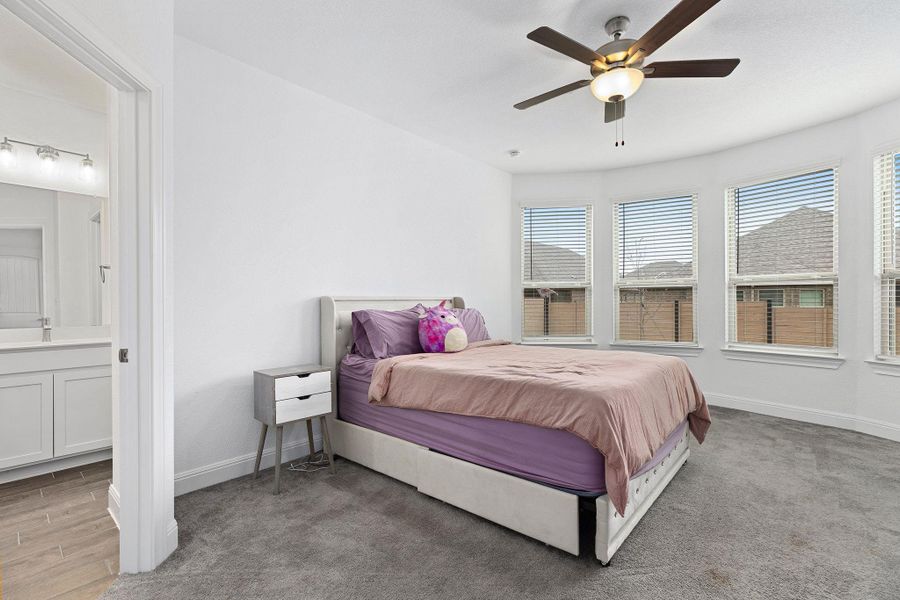A well-lit primary bedroom features soft carpeting, bay windows, and a ceiling fan.