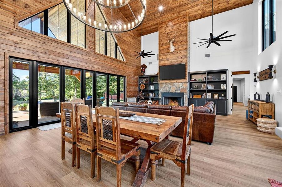 Dining area with a towering ceiling, a stone fireplace, light wood finished floors, wood walls, and ceiling fan Dining area with a towering ceiling, a stone fireplace, light wood finished floors, wood walls, and ceiling fan