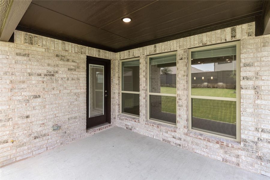 Covered back patio with brick exterior, dark wood ceiling, back door, and large windows overlooking a grassy yard Covered back patio with brick exterior, dark wood ceiling, back door, and large windows overlooking a grassy yard