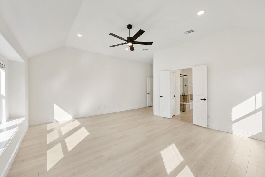 Empty bedroom with vaulted ceiling, black ceiling fan, light wood floors, and open ensuite bathroom door Empty bedroom with vaulted ceiling, black ceiling fan, light wood floors, and open ensuite bathroom door