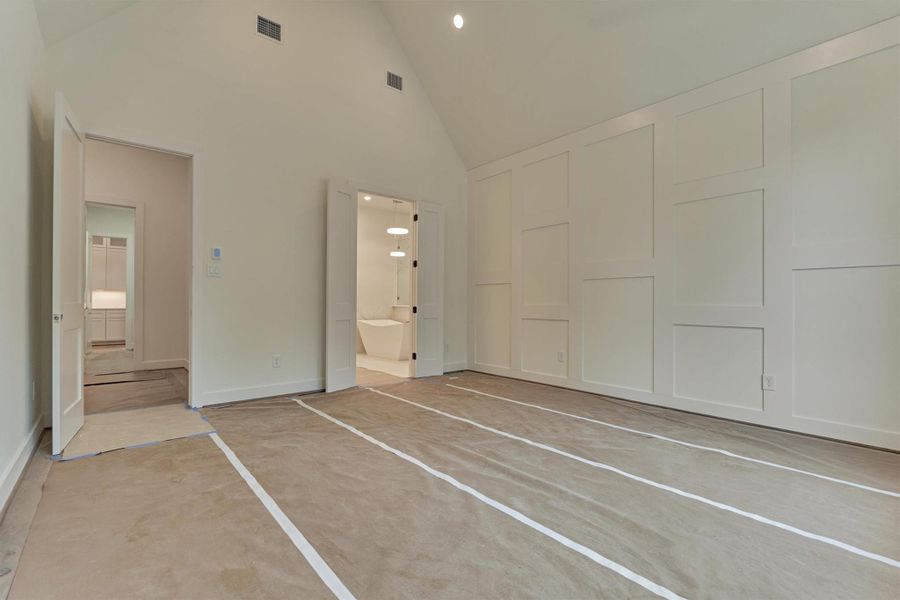 Spacious primary suite with vaulted ceilings and custom wall paneling, opening to a luxurious en-suite bathroom through elegant double doors.