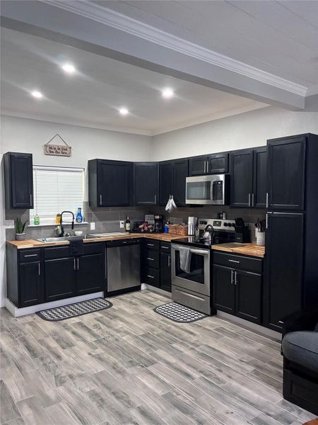 Kitchen with dark cabinetry, butcher counter tops, crown molding, appliances with stainless steel finishes, tasteful backsplash, and tile floors