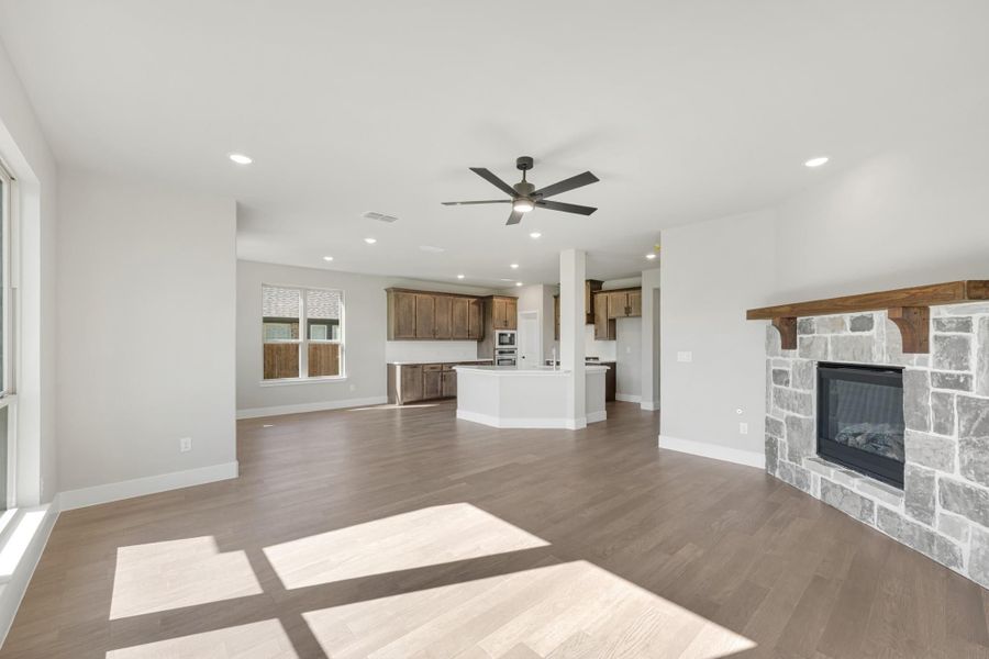 Open living area with stone fireplace, hardwood floors, ceiling fan, and open kitchen with dark wood cabinets in background