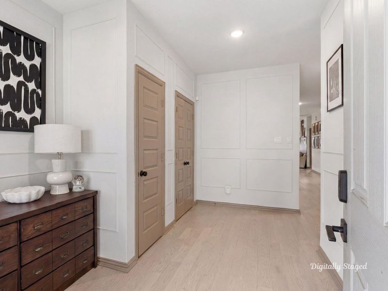 Bright entry foyer with wide-plank wood flooring and detailed picture-frame wall molding that adds a custom, polished touch. A built-in coat closet and open sightline lead naturally into the main living area. This photo has been digitally staged to illustrate the possibilities of the home's aesthetic and potential design. Some furnishings and décor shown are for visual inspiration.