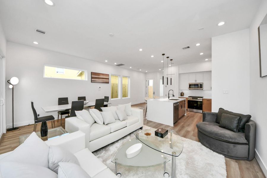 This modern, open-concept living room seamlessly blends into a sleek kitchen, featuring elegant white walls, warm wood floors, and abundant natural light, creating a bright and inviting space perfect for relaxation and entertaining.