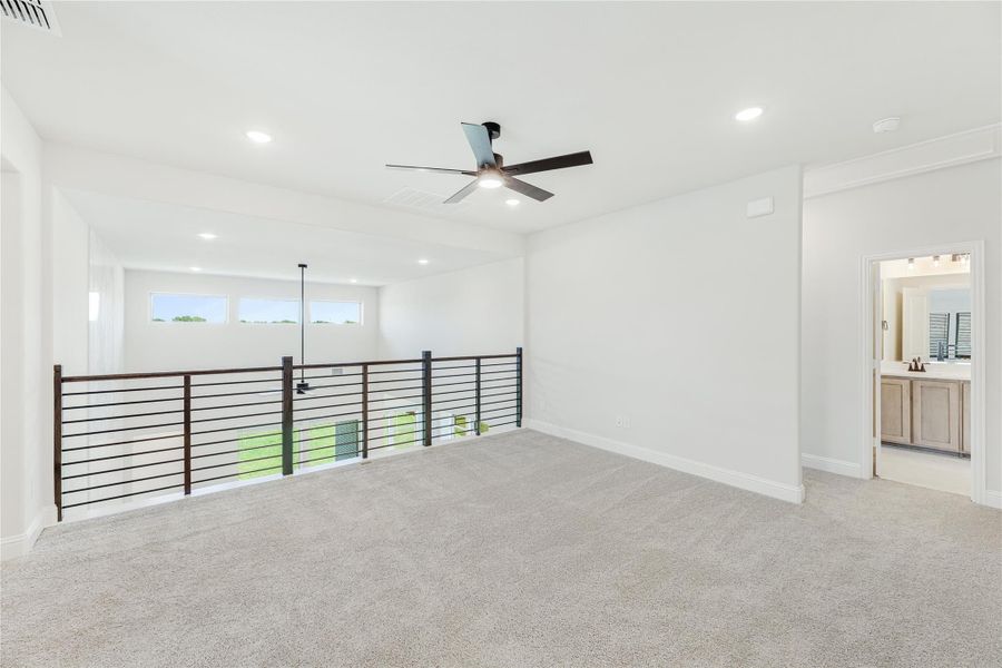 Open upstairs loft with carpet, ceiling fan, metal railing overlooking lower level, and adjoining bathroom doorway.