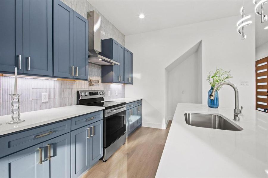 Kitchen with stainless steel range with electric cooktop, blue cabinets, wall chimney range hood, light stone counters, and recessed lighting