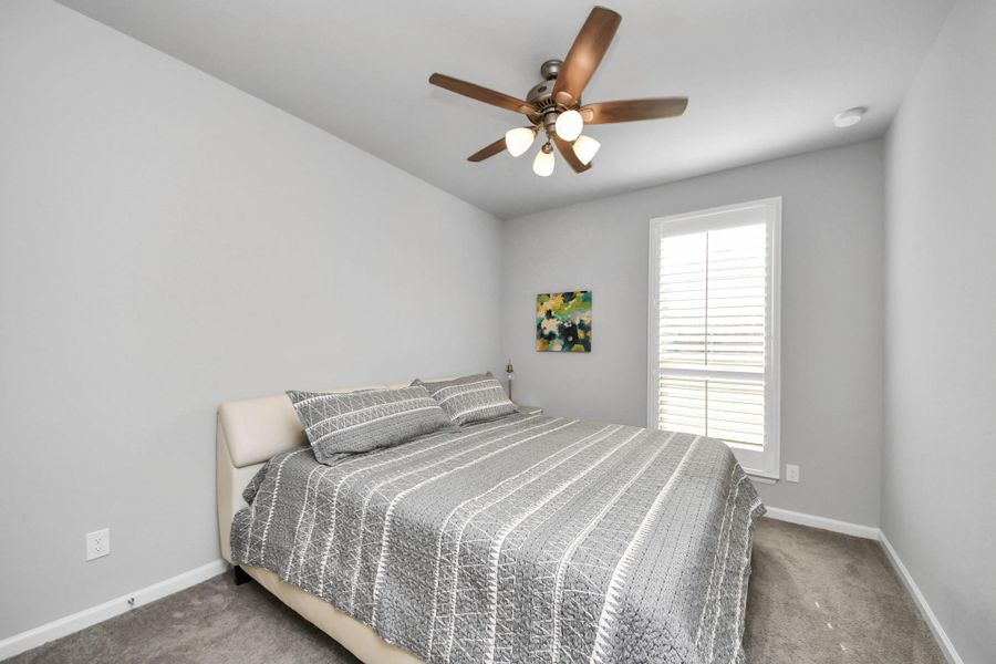 This bedroom features a cozy setup with a large bed, gray walls, and carpeted flooring. It includes a ceiling fan for comfort and a window with shutters allowing natural light. This bedroom features a cozy setup with a large bed, gray walls, and carpeted flooring. It includes a ceiling fan for comfort and a window with shutters allowing natural light.
