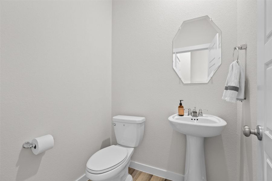 Modern, minimalist bathroom featuring a pedestal sink, toilet, and octagonal mirror. Neutral tones with a light wood floor create a clean, stylish look.