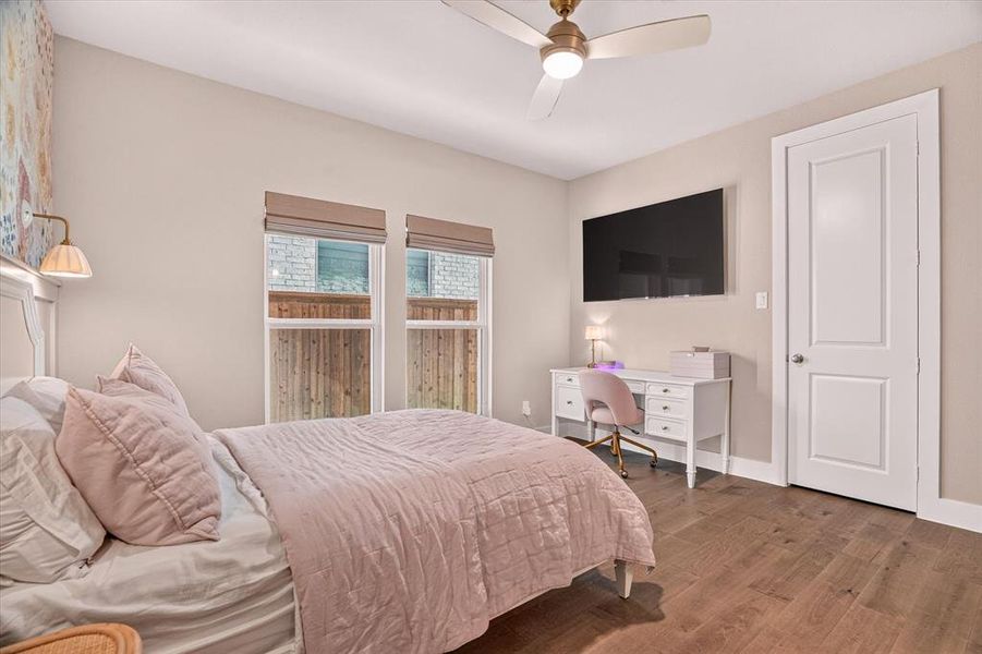 Bedroom featuring wood-finish flooring, two large windows with privacy blinds, a ceiling fan with integrated lighting, and a paneled interior door Bedroom featuring wood-finish flooring, two large windows with privacy blinds, a ceiling fan with integrated lighting, and a paneled interior door