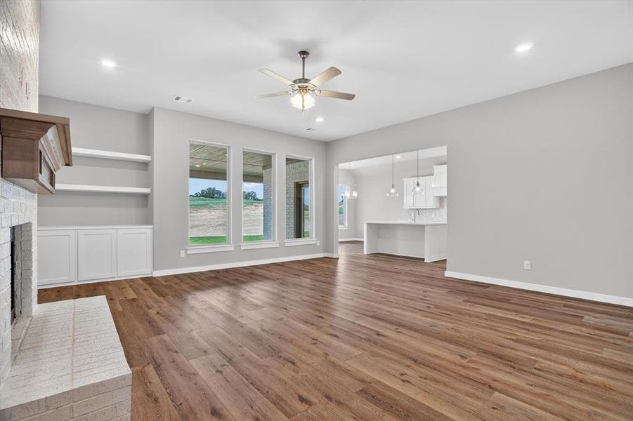 Spacious interior with wood-finish flooring, a white brick fireplace with a wood mantle, and built-in shelving with cabinetry Spacious interior with wood-finish flooring, a white brick fireplace with a wood mantle, and built-in shelving with cabinetry