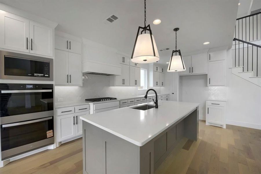 Contemporary kitchen featuring a large central island with a solid surface countertop, white shaker cabinetry with black hardware, stainless steel double ovens and microwave, a gas range with a custom vent hood, and wood-finish flooring Contemporary kitchen featuring a large central island with a solid surface countertop, white shaker cabinetry with black hardware, stainless steel double ovens and microwave, a gas range with a custom vent hood, and wood-finish flooring