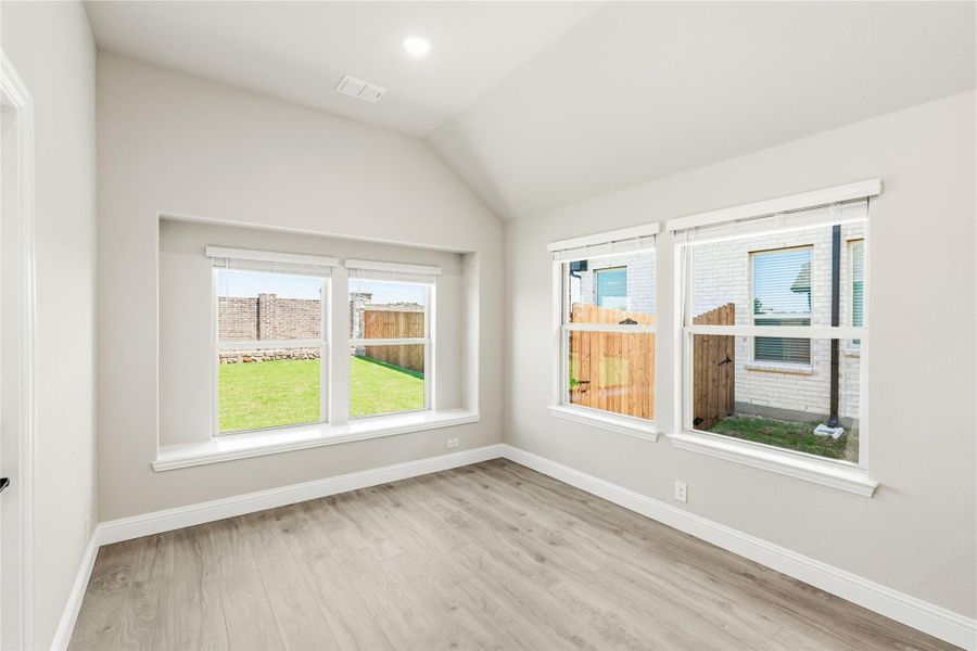 Empty room with vaulted ceiling, light wood floors, and three windows overlooking a fenced backyard