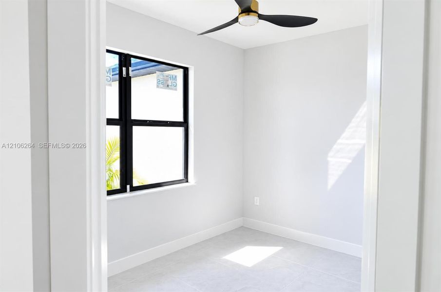 Neutral-toned room featuring a matte black three-blade ceiling fan with gold accents, large-format tile flooring, and a single-hung window with a matte black frame