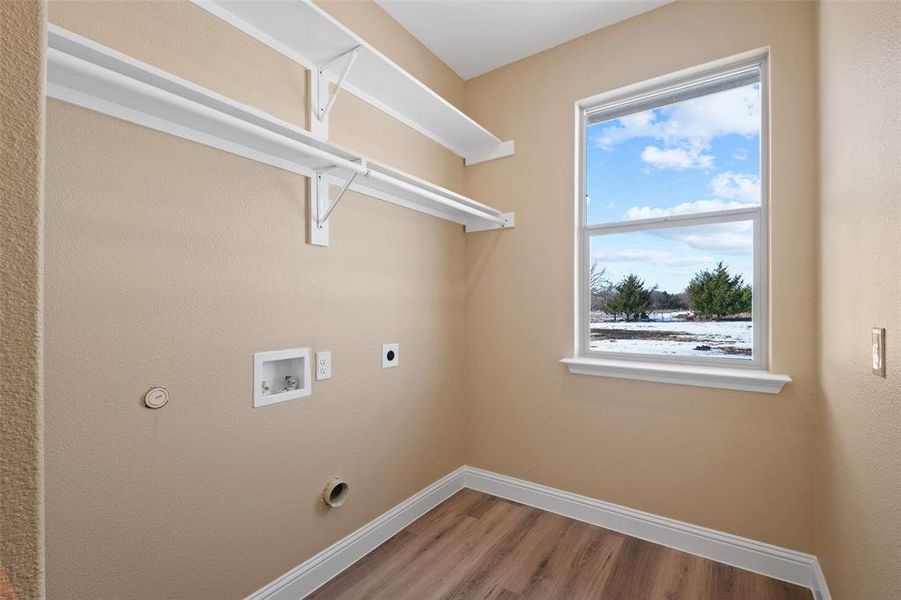 Utility room featuring a large window, wood-finish flooring, white baseboards, and built-in white shelving with a hanging rod Utility room featuring a large window, wood-finish flooring, white baseboards, and built-in white shelving with a hanging rod