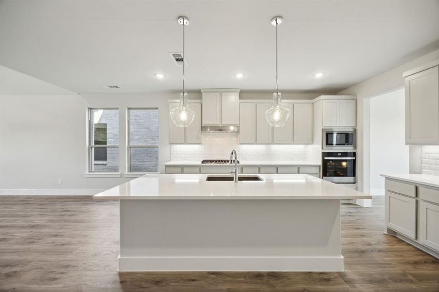 Kitchen with backsplash, dark wood-type flooring, an island with sink, decorative light fixtures, and stainless steel appliances