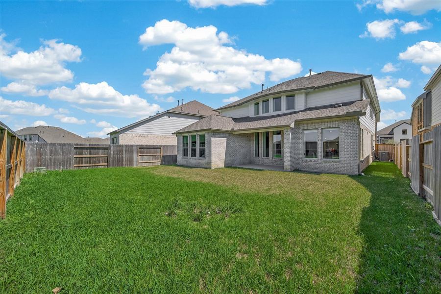 The photo shows a spacious backyard with well-maintained grass, enclosed by wooden fencing. The house is a two-story brick structure with large windows and a patio, ideal for outdoor activities and relaxation.