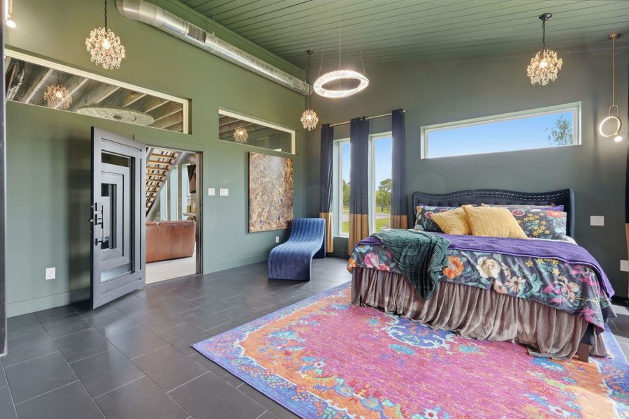 This spacious bedroom features modern industrial design with exposed ductwork, elegant lighting, and large windows for natural light. The vibrant decor includes a colorful area rug and a plush, floral bedspread, creating a warm and inviting atmosphere.