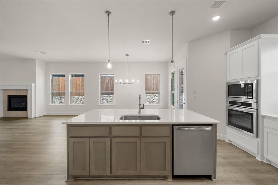 Kitchen with stainless steel appliances, light countertops, light wood-type flooring, a center island with sink, and a brick fireplace
